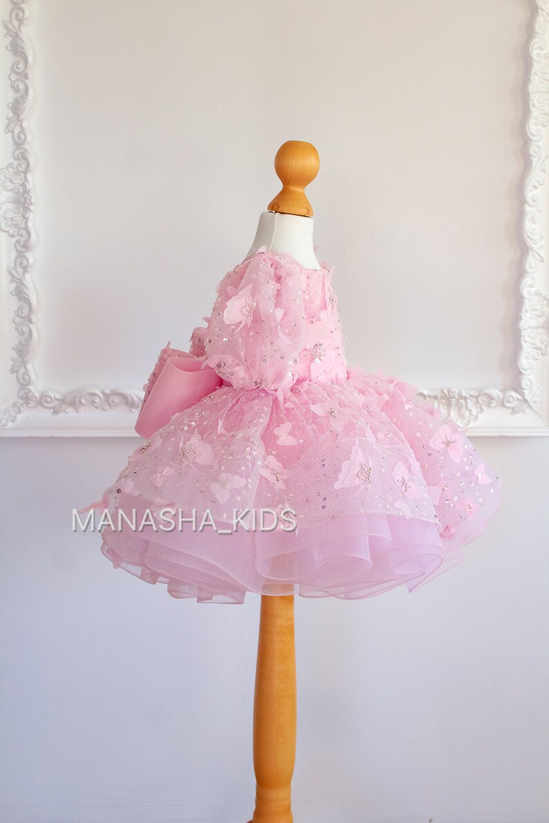 May include: A pink tulle dress with butterfly embellishments and a large pink bow. The dress is on a wooden mannequin.