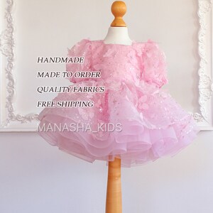 May include: A pink tulle dress with butterfly embellishments and sequins. The dress is on a wooden mannequin. The text "HANDMADE", "MADE TO ORDER", "QUALITY FABRICS", "FREE SHIPPING", and "MANASHA_KIDS" is printed on the image.