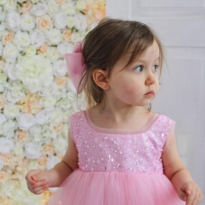 May include: A young girl wearing a pink tulle dress with a sequined bodice. The dress has a round neckline and sleeveless design. The girl is standing in front of a wall of white and pink flowers. The text "MANASHA_KIDS" is visible at the bottom of the image.