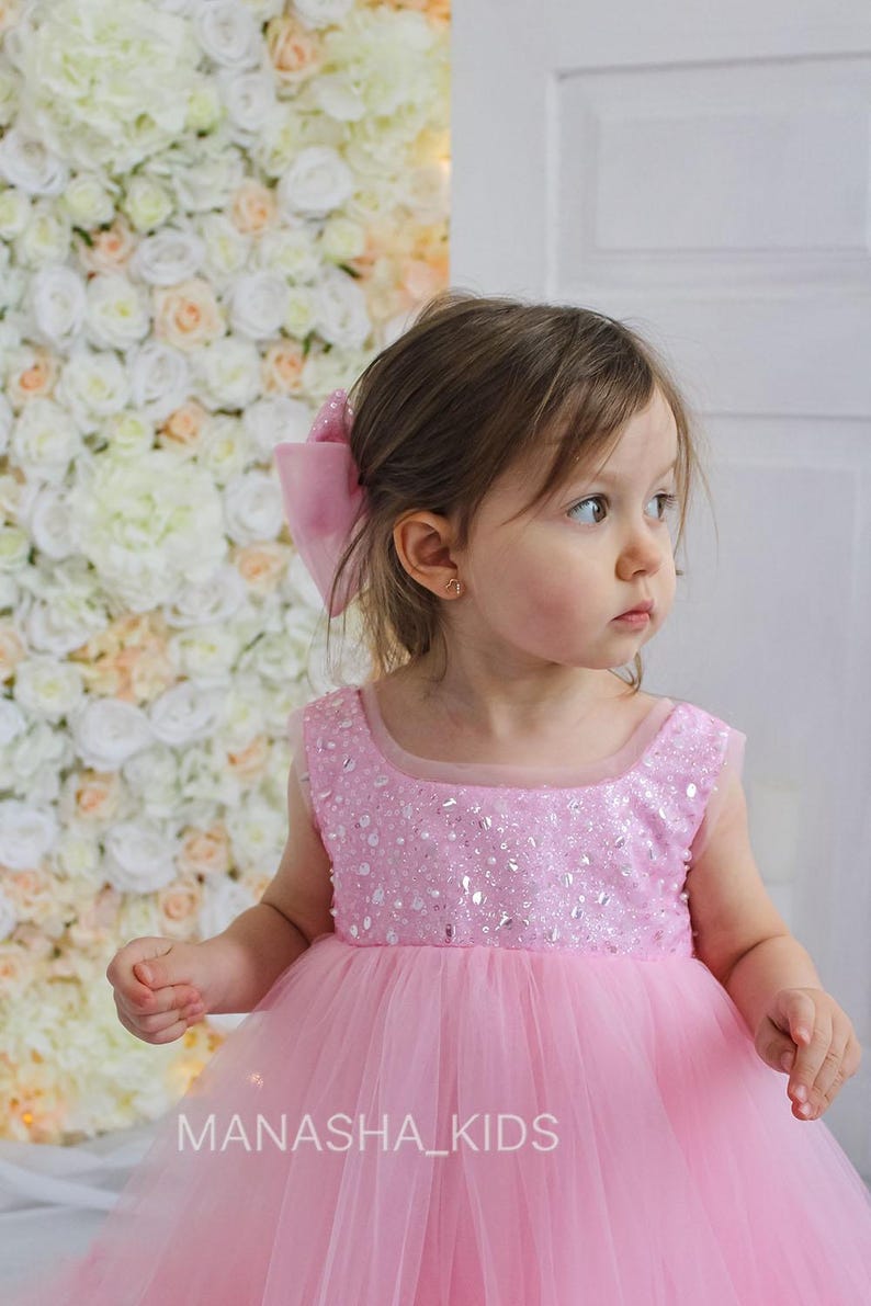 May include: A young girl wearing a pink tulle dress with a sequined bodice. The dress has a round neckline and sleeveless design. The girl is standing in front of a wall of white and pink flowers. The text "MANASHA_KIDS" is visible at the bottom of the image.