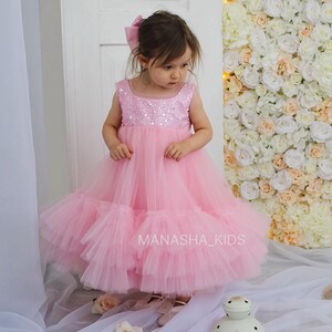 May include: A little girl wearing a pink tulle dress with sequins. The dress has multiple layers of tulle and a sparkly bodice. The girl is standing in front of a wall of white and pink flowers. The text "MANASHA_KIDS" is visible on the dress.