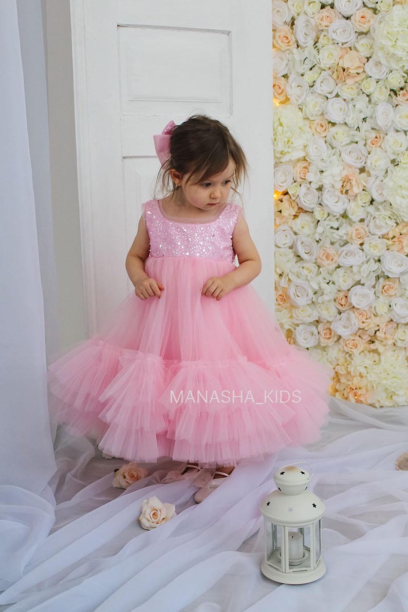May include: A little girl wearing a pink tulle dress with sequins. The dress has multiple layers of tulle and a sparkly bodice. The girl is standing in front of a wall of white and pink flowers. The text "MANASHA_KIDS" is visible on the dress.