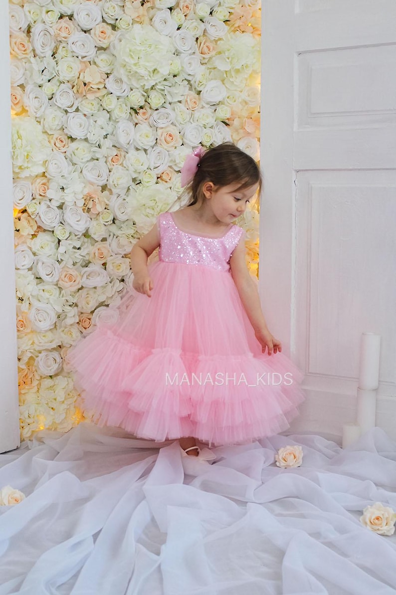 May include: A young girl wearing a pink tulle dress with sequins. The dress has multiple layers of tulle and a sequined bodice. The girl is standing in front of a white flower wall.