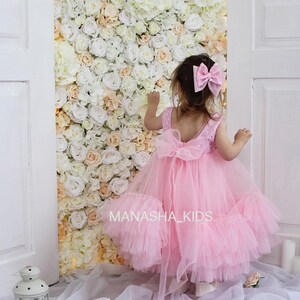 May include: A little girl wearing a pink tulle dress with a sequined bodice and a large pink bow in her hair. The dress has multiple layers of tulle and a bow at the back. The girl is standing in front of a wall of white and peach artificial flowers.