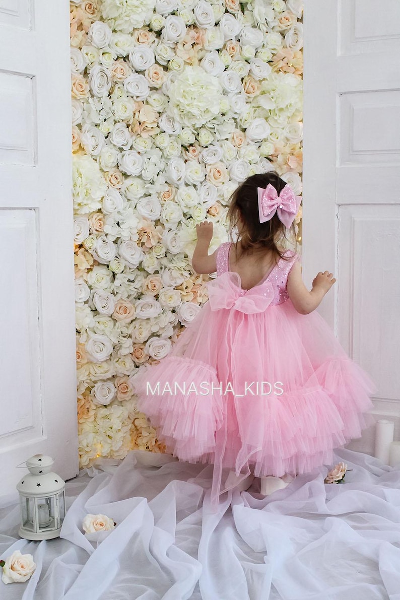 May include: A little girl wearing a pink tulle dress with a sequined bodice and a large pink bow in her hair. The dress has multiple layers of tulle and a bow at the back. The girl is standing in front of a wall of white and peach artificial flowers.