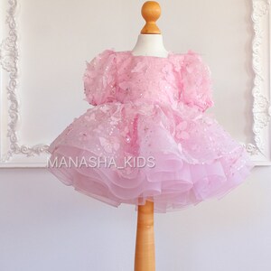 May include: A pink tulle dress with butterfly appliques and sequins. The dress is on a wooden mannequin and has a layered skirt.