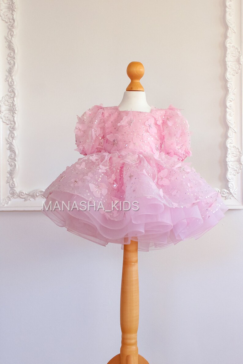 May include: A pink tulle dress with butterfly appliques and sequins. The dress is on a wooden mannequin and has a layered skirt.