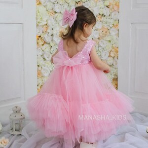 May include: A little girl wearing a pink tulle dress with sequins and a large pink bow in her hair. The dress has a ruffled skirt and a bow on the back. The girl is standing in front of a wall of white flowers.