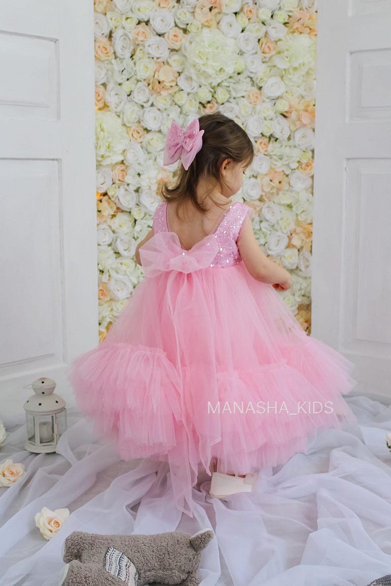 May include: A little girl wearing a pink tulle dress with sequins and a large pink bow in her hair. The dress has a ruffled skirt and a bow on the back. The girl is standing in front of a wall of white flowers.
