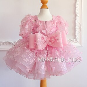 May include: A pink tulle dress with butterfly appliques and a large pink bow. The dress is on a mannequin and has the text "MANASHA_KIDS" on the bottom.