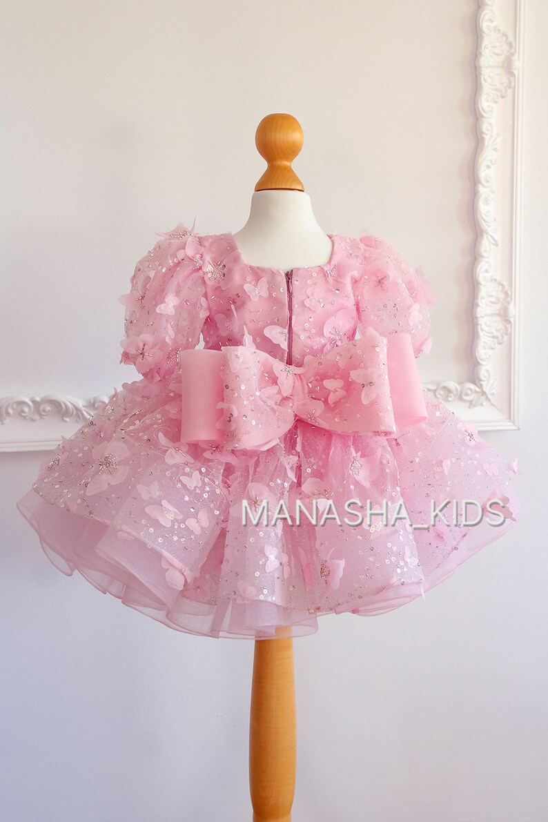 May include: A pink tulle dress with butterfly appliques and a large pink bow. The dress is on a mannequin and has the text "MANASHA_KIDS" on the bottom.