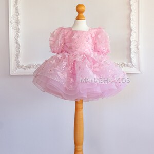 May include: A pink tulle dress with butterfly and star embellishments. The dress is on a wooden dress form. The text "MANASHA_KIDS" is visible on the dress.