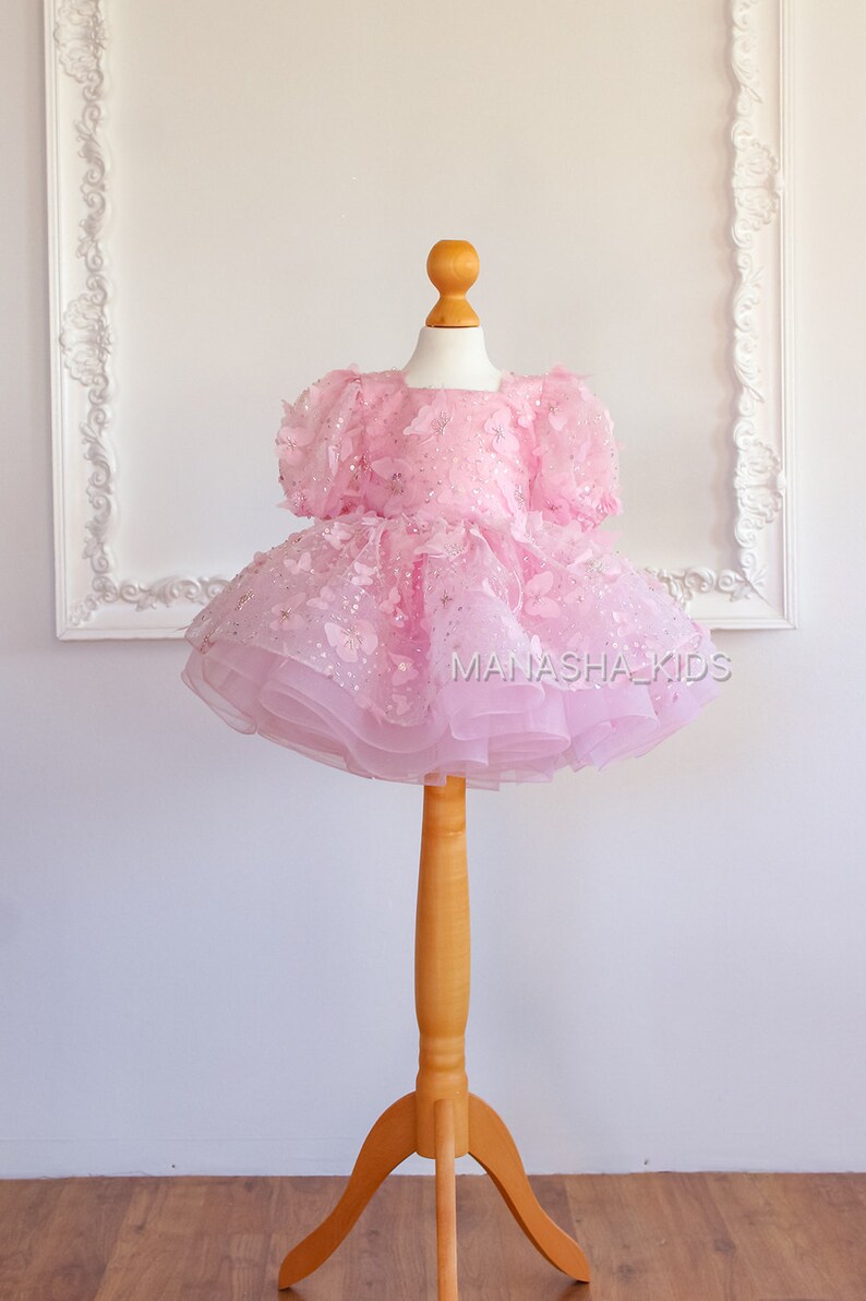 May include: A pink tulle dress with butterfly and star embellishments. The dress is on a wooden dress form. The text "MANASHA_KIDS" is visible on the dress.