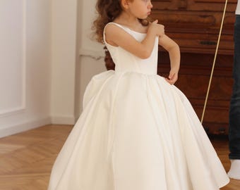 May include: A young girl in a white satin dress with a full skirt and small train. The dress has a simple bodice and a large bow in the back. She wears black shoes with gold chain detailing. The background includes a wooden piano.