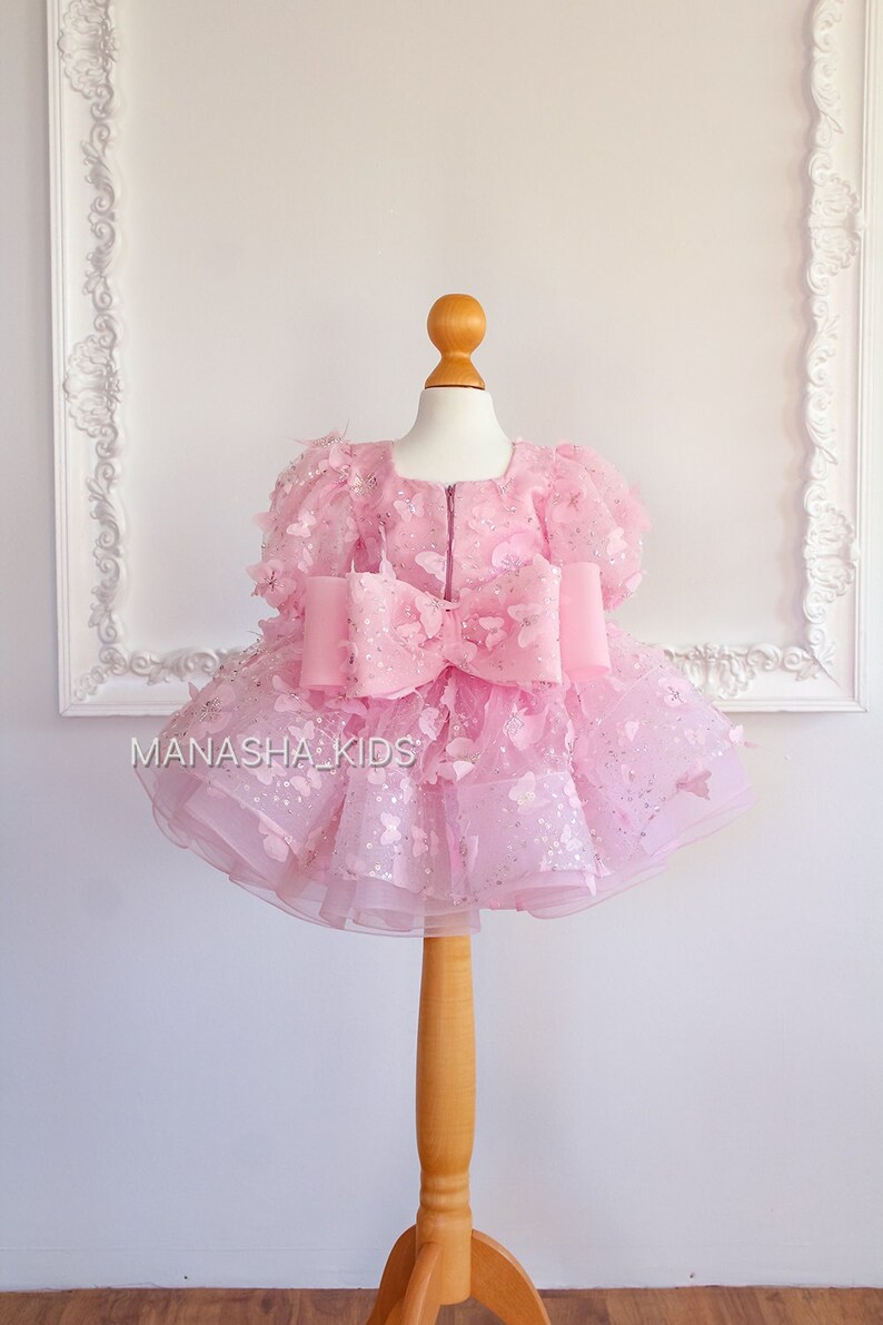 May include: A pink tulle dress with butterfly appliques and a large pink bow. The dress is on a wooden mannequin.