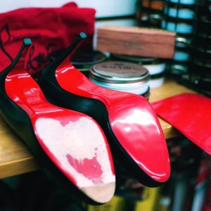 May include: Two pairs of black high-heeled shoes with red soles. The shoes are on a wooden surface and there is a red bag in the background.