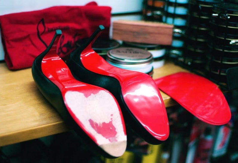 May include: Two pairs of black high-heeled shoes with red soles. The shoes are on a wooden surface and there is a red bag in the background.