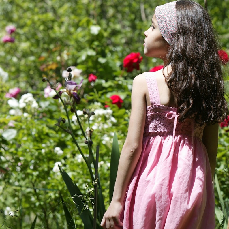 Petite fille de dos dans un jardin fleuri, avec une robe à bretelles coloris fraise, laçée dans le dos.