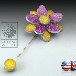 Puede incluir: Un broche de flor amarillo y morado con un alfiler de plata. La flor tiene una superficie texturizada y está hecha de metal.