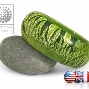 Puede incluir: Un anillo verde con un patrón de mármol blanco y verde. El anillo está apoyado en una piedra gris.