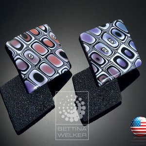 Pode incluir: Dois brincos quadrados com um padrão geométrico em preto, branco e roxo. Os brincos estão sobre uma superfície preta texturizada.