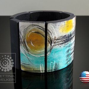 May include: A wide black and white bracelet with a colorful abstract design. The design features shades of blue, yellow, and white. The bracelet is made of multiple pieces of wood that are joined together.
