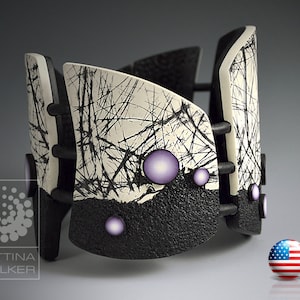 Pode incluir: Um bracelete de cerâmica preto e branco com uma superfície texturizada e detalhes roxos. O bracelete é feito de várias peças que são unidas por conectores metálicos pretos.