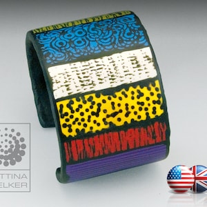 Könnte beinhalten: Ein breites, schwarzes Manschettenarmband mit einem bunten geometrischen Muster. Das Muster enthält blaue, weiße, gelbe, rote und violette Streifen. Das Armband ist aus Polymer Clay.