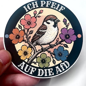 Puede incluir: Una pegatina circular con un gorrión marrón y blanco posado en una rama rodeado de flores coloridas en tonos rosa, naranja, amarillo, verde y azul. El texto ICH PFEIF AUF DIE AFD está impreso en blanco a lo largo del borde negro.