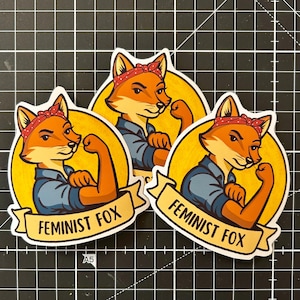 Könnte beinhalten: Drei gelbe und weiße runde Aufkleber mit einem Comic-Fuchs, der ein rotes Bandana und ein blaues Hemd trägt. Der Fuchs beugt seinen Bizeps und der Text "Feminist Fox" befindet sich auf einem Banner unter dem Fuchs.