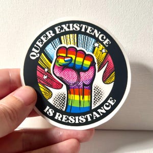 Op de afbeelding: Een ronde sticker met een zwarte achtergrond en witte tekst die "Queer Existence is Resistance" luidt. In het midden van de sticker bevindt zich een regenboogvuist.
