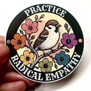 Könnte beinhalten: Ein runder Aufkleber mit einem braun-weißen Spatzen, der auf einem Ast sitzt und von bunten Blumen in Rosa, Orange, Gelb, Grün, Blau und Lila umgeben ist. Der Text "PRACTICE RADICAL EMPATHY" ist in Weiß auf einem schwarzen Rand gedruckt.