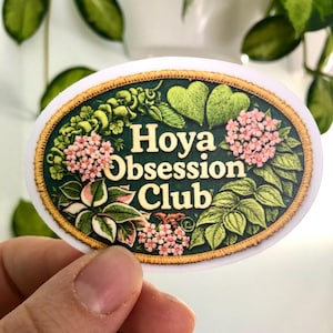 May include: An oval sticker featuring the text "Hoya Obsession Club" with botanical illustrations in green, pink, and white. The sticker has a gold border and is held by a hand.