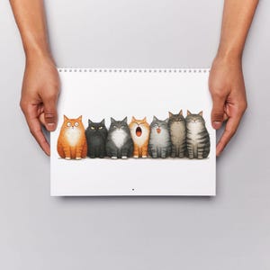 May include: A spiral-bound notepad featuring a row of seven cartoon cats with various expressions. The cats are in shades of orange, black, gray, and brown. The notepad is held by two hands against a white background.