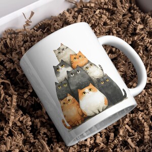 May include: White ceramic mug featuring a watercolor illustration of a group of cats in various colors, including orange, gray, and black. The cats are stacked together, creating a playful design. The mug has a white handle and is set against a brown background.