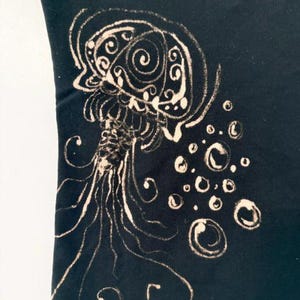 May include: Black t-shirt featuring a jellyfish design. The jellyfish is depicted in a cream color with intricate details, including swirling patterns and flowing tentacles. Bubbles are also included in the design.