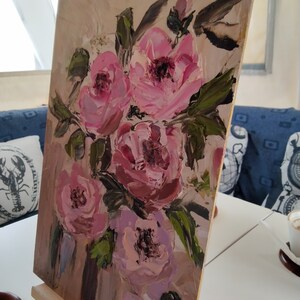 Puede incluir: Una pintura de rosas rosas con hojas verdes sobre un lienzo. La pintura est&aacute; en un caballete y se apoya contra una pared.