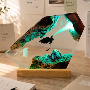 Könnte beinhalten: Dekorative Harzlampe mit einer Fantasy-Szene mit zwei Drachen. Die Lampe hat einen Holzsockel und einen klaren, abgewinkelten Harzkörper. Im Inneren sind ein schwarzer und ein weißer Drache dargestellt, mit einem türkisfarbenen Leuchten.