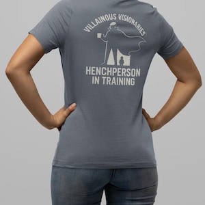 Puede incluir: Camiseta azul grisáceo con el texto blanco « VILLAINOUS VISIONARIES » y « HENCHPERSON IN TRAINING ». Un dibujo de una persona con capa sosteniendo una taza también está impreso en la espalda. La persona lleva vaqueros azules.