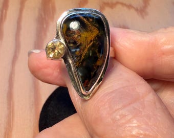 May include: A silver ring featuring a large, teardrop-shaped dark stone with golden-brown inclusions and a smaller, round, faceted yellow gemstone set to the side. The metal band is polished and frames both stones in a smooth, bezel-style setting.