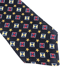 May include: A dark navy blue necktie featuring a repeating pattern of small gold five-pointed stars and square geometric icons. Each square icon is divided into four triangles colored in red, blue, and yellow, arranged in a consistent grid.