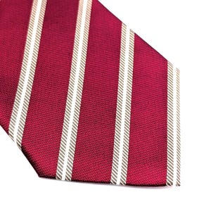May include: A necktie featuring a deep burgundy red base color with a repeating pattern of vertical white stripes. The fabric has a visible diagonal weave texture throughout the surface, and the tie ends in a pointed tip at the bottom edge.