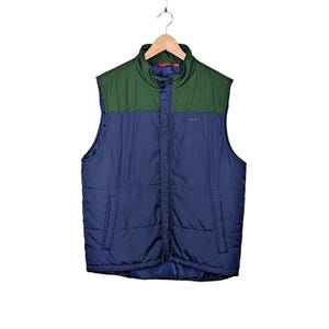 May include: A quilted puffer vest featuring a dark blue lower section and a forest green upper yoke. The vest has a stand-up collar, a central snap-button placket, and two side pockets. The word TAUD is embroidered in small letters on the chest.