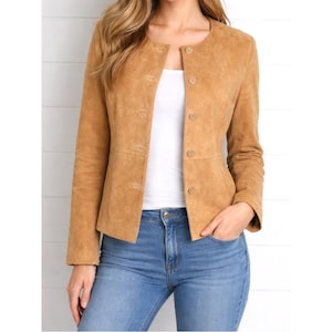 May include: A tan suede jacket featuring a collarless round neckline, long sleeves, and a front button closure with five matching tan buttons. The jacket is worn over a plain white top and paired with blue denim jeans, creating a layered look.