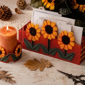 May include: A plastic canvas desk organiser and a matching candle holder, both in a vibrant orange hue. The organiser features three stitched sunflower motifs with dark brown centres and yellow petals, while the candle holder has two.