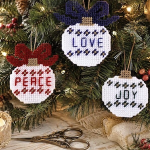 May include: Three white plastic canvas ornaments featuring stitched designs. One ornament has a red bow and the word PEACE, one has a dark blue bow and the word LOVE, and one has a black bow and the word JOY. Each has a gold-colored cap and string.