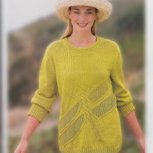 Può includere: Una donna indossa un maglione di lana giallo con un motivo geometrico e un cappello di paglia. È in piedi davanti a uno sfondo sfocato.