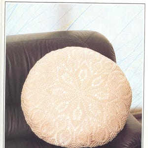 Może przedstawiać: Okrągła poduszka wykonana na szydełku z delikatnym wzorem kwiatowym w kremowym kolorze. Poduszka leży na brązowej skórzanej sofie.