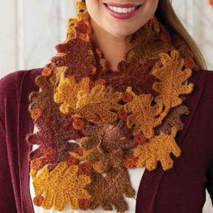 Op de afbeelding: Een vrouw draagt een bordeauxrode cardigan over een wit shirt en een gehaakte sjaal gemaakt van bruine, oranje en gele bladeren. De sjaal is een herfst-accessoire.