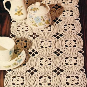 May include: A white crocheted table runner with a square pattern. The runner is laid on a wooden table with a cream colored creamer, sugar bowl, and two teacups and saucers.
